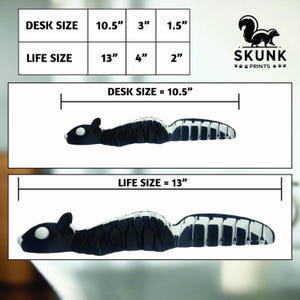 May include: A black and white skunk-shaped desk accessory. The image shows two sizes: desk size at 10.5 inches long and life size at 13 inches long. The Skunk Prints logo is in the upper right corner.
