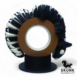May include: A decorative tape dispenser featuring a skunk design. The skunk is black and white, with a brown tape roll in the center. The dispenser sits on a black base, with the "SKUNK PRINTS" logo visible.