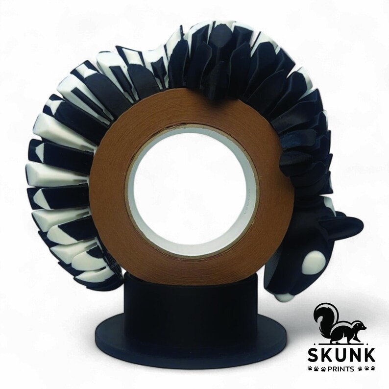 May include: A decorative tape dispenser featuring a skunk design. The skunk is black and white, with a brown tape roll in the center. The dispenser sits on a black base, with the "SKUNK PRINTS" logo visible.