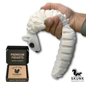 May include: A white articulated squirrel fidget toy held in a hand. The toy is made of interlocking segments. A black box with the text "PREMIUM FIDGETS" and "CALM THE CHAOS" is in the lower left. The Skunk Prints logo is in the lower right.