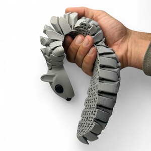 May include: A gray, articulated squirrel toy held in a hand. The toy is made of interlocking segments, with a detailed, textured design. The squirrel's head has a black eye and small ears. The toy is flexible and can be posed.