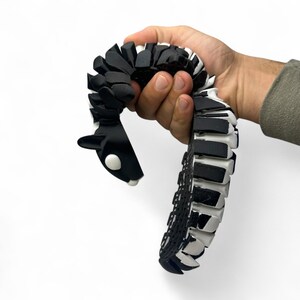 May include: A black and white articulated snake toy with a rounded head and a segmented body. The toy is held in a hand against a white background. The snake's head has a white eye and a white spot on the chin.