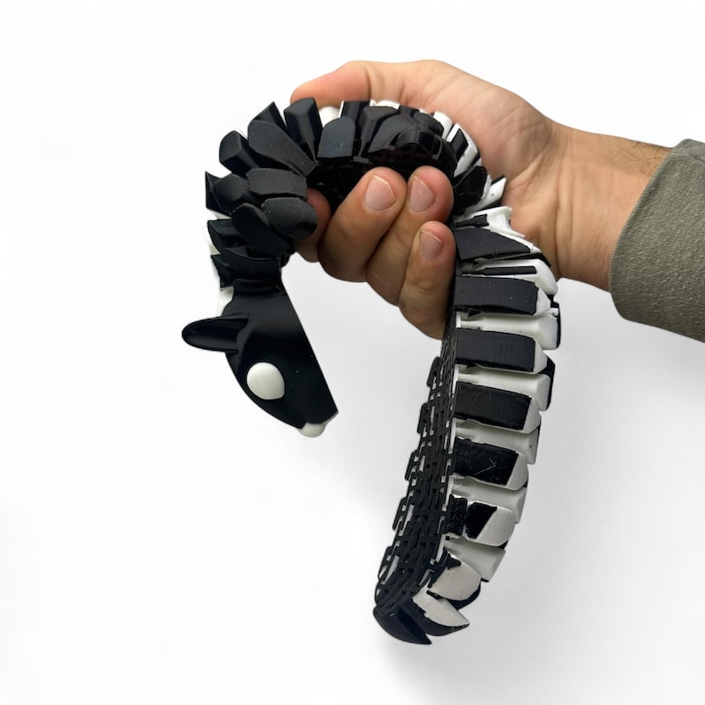 May include: A black and white articulated snake toy with a rounded head and a segmented body. The toy is held in a hand against a white background. The snake's head has a white eye and a white spot on the chin.