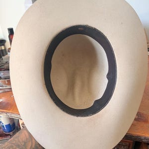 K&ouml;nnte beinhalten: Hellbeige Cowboyhut mit schwarzem Innenband. Der Hut wird von innen betrachtet, wobei die Form der Krone und die N&auml;hte des Bands zu sehen sind. Der Hut scheint aus Filz oder einem &auml;hnlichen Material zu bestehen.