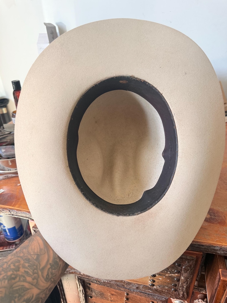 K&ouml;nnte beinhalten: Hellbeige Cowboyhut mit schwarzem Innenband. Der Hut wird von innen betrachtet, wobei die Form der Krone und die N&auml;hte des Bands zu sehen sind. Der Hut scheint aus Filz oder einem &auml;hnlichen Material zu bestehen.