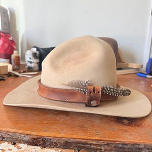 K&ouml;nnte beinhalten: Beigefarbener Fedora-Hut mit braunem Lederband und Federakzenten. Der Hut hat eine breite Krempe und eine leicht gebogene Krone. Das Lederband ist mit einer kleinen Schnalle befestigt. Die Federn sind braun und wei&szlig;.
