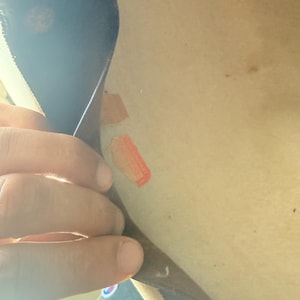 K&ouml;nnte beinhalten: Eine Hand h&auml;lt ein dunkles Objekt, m&ouml;glicherweise Leder oder Stoff, mit zwei kleinen, rechteckigen, orangefarbenen und beigefarbenen Aufklebern auf einer hellen Oberfl&auml;che. Der Hintergrund ist unscharf.