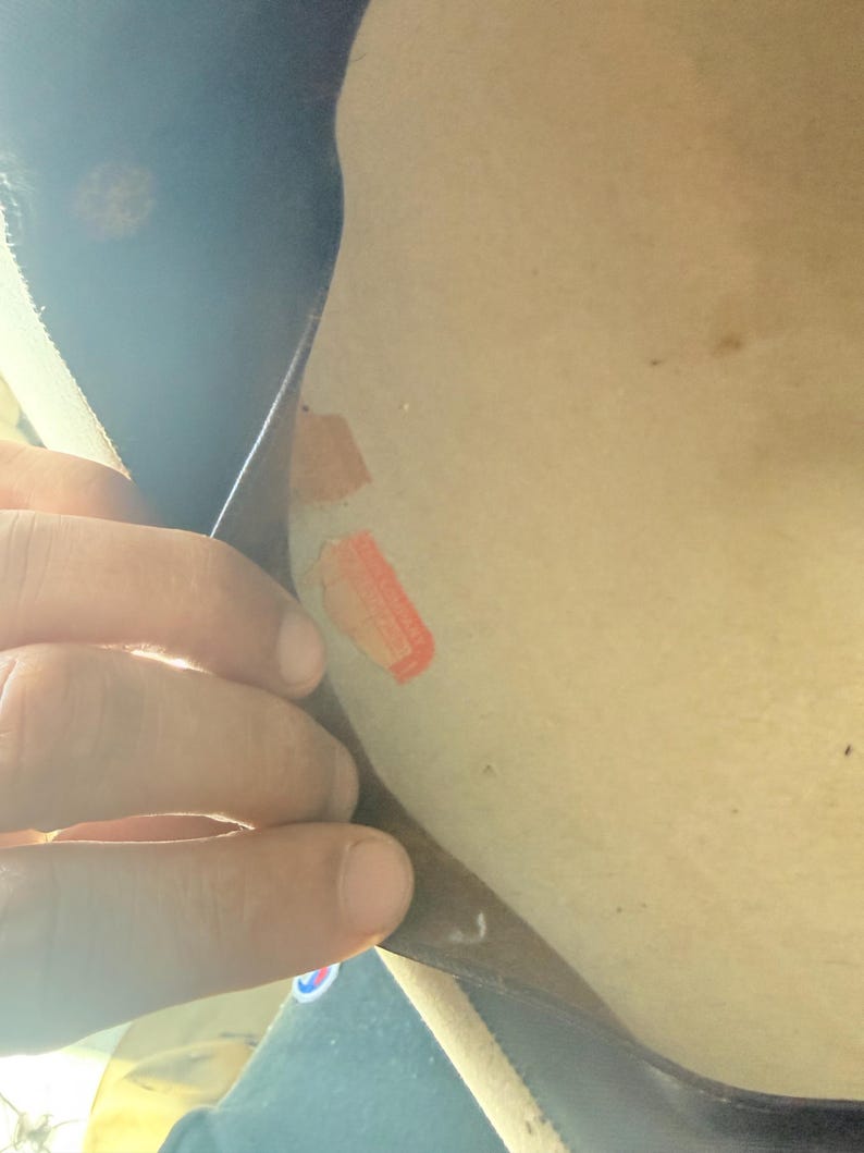 K&ouml;nnte beinhalten: Eine Hand h&auml;lt ein dunkles Objekt, m&ouml;glicherweise Leder oder Stoff, mit zwei kleinen, rechteckigen, orangefarbenen und beigefarbenen Aufklebern auf einer hellen Oberfl&auml;che. Der Hintergrund ist unscharf.