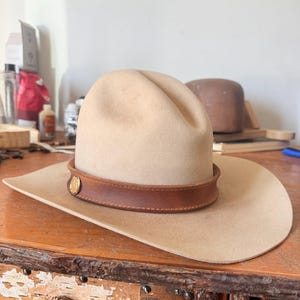 K&ouml;nnte beinhalten: Beigefarbener Cowboyhut mit braunem Lederband und goldenem Emblem. Der Hut hat eine breite Krempe und eine abgerundete Krone. Der Hut liegt auf einer Holzoberfl&auml;che.