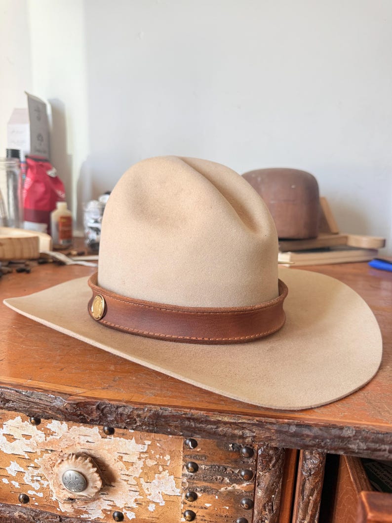K&ouml;nnte beinhalten: Beigefarbener Cowboyhut mit braunem Lederband und goldenem Emblem. Der Hut hat eine breite Krempe und eine abgerundete Krone. Der Hut liegt auf einer Holzoberfl&auml;che.