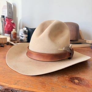 K&ouml;nnte beinhalten: Ein beigefarbener Cowboyhut mit einem braunen Lederband und einer dekorativen Feder. Der Hut liegt auf einer Holzoberfl&auml;che. Der Hut hat eine breite Krempe und eine abgerundete Krone.