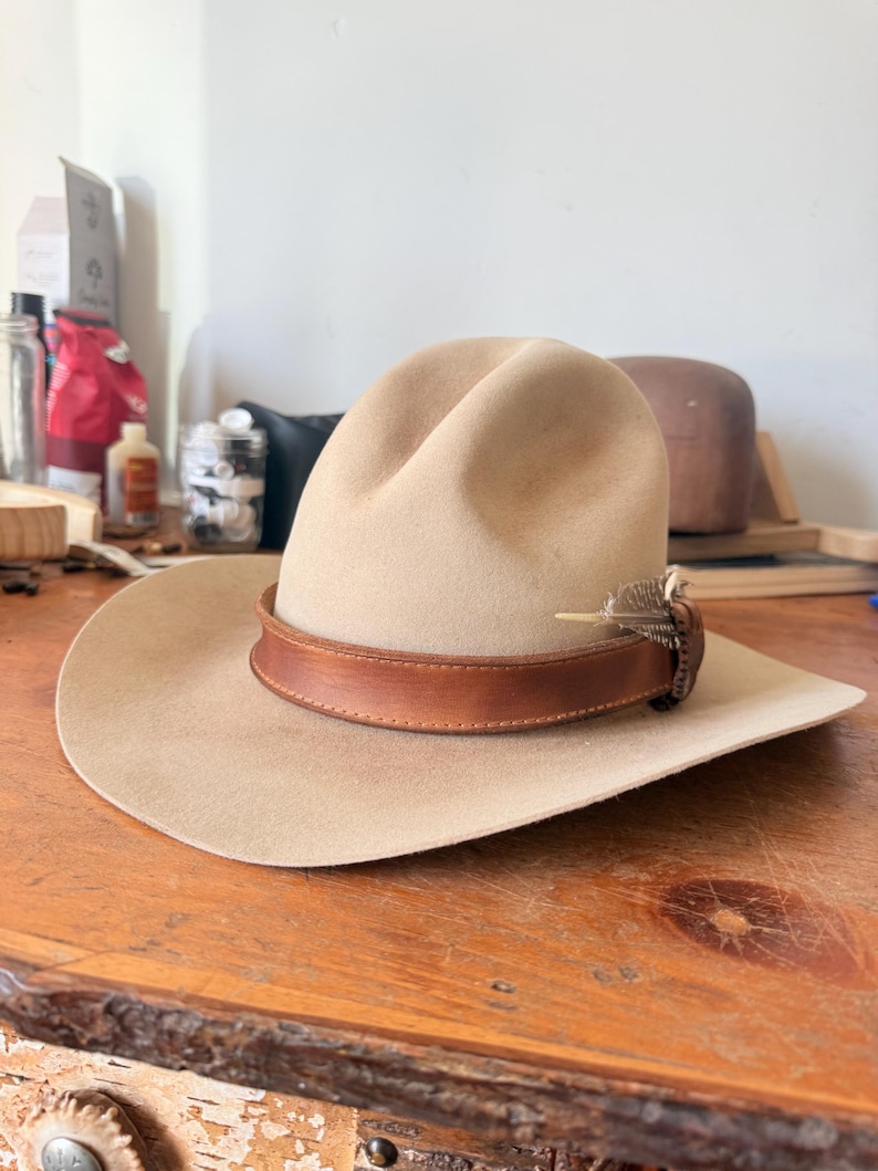 K&ouml;nnte beinhalten: Ein beigefarbener Cowboyhut mit einem braunen Lederband und einer dekorativen Feder. Der Hut liegt auf einer Holzoberfl&auml;che. Der Hut hat eine breite Krempe und eine abgerundete Krone.