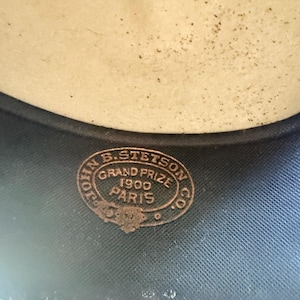 K&ouml;nnte beinhalten: Nahaufnahme des Inneren eines Hutes. Der Hut hat eine hellbeige Krone und eine dunkelblaue Krempe. Ein kreisf&ouml;rmiges Bronzeemblem lautet "JOHN B. STETSON CO. GRAND PRIZE 1900 PARIS."