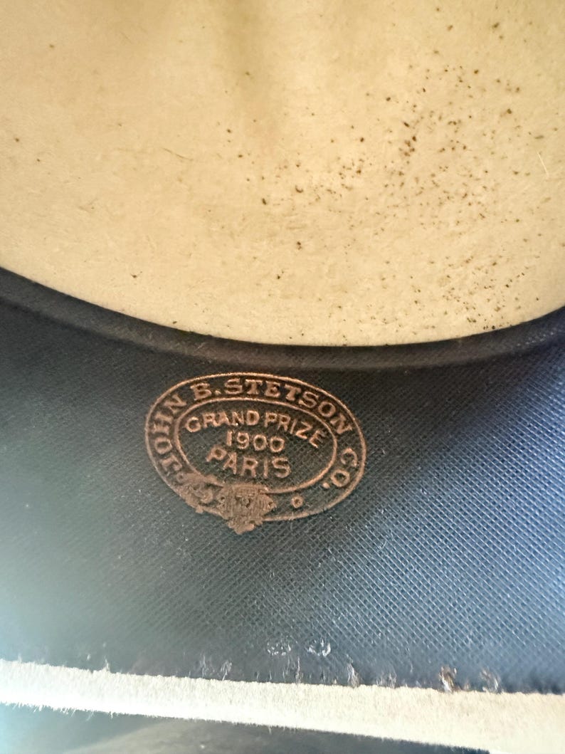 K&ouml;nnte beinhalten: Nahaufnahme des Inneren eines Hutes. Der Hut hat eine hellbeige Krone und eine dunkelblaue Krempe. Ein kreisf&ouml;rmiges Bronzeemblem lautet "JOHN B. STETSON CO. GRAND PRIZE 1900 PARIS."