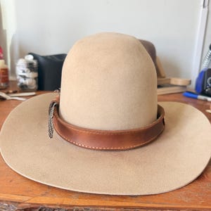 K&ouml;nnte beinhalten: Beigefarbener Cowboyhut mit braunem Lederband und silberner Kette. Der Hut hat eine breite Krempe und eine abgerundete Krone. Der Hut liegt auf einer Holzoberfl&auml;che.