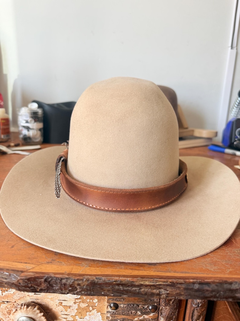 K&ouml;nnte beinhalten: Beigefarbener Cowboyhut mit braunem Lederband und silberner Kette. Der Hut hat eine breite Krempe und eine abgerundete Krone. Der Hut liegt auf einer Holzoberfl&auml;che.