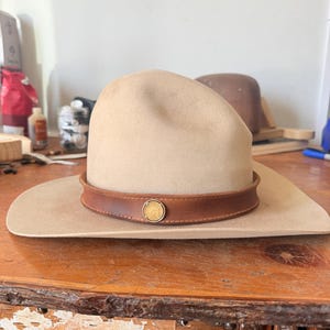 K&ouml;nnte beinhalten: Ein beigefarbener Cowboyhut mit einem braunen Lederband und einem goldfarbenen kreisf&ouml;rmigen Emblem. Der Hut hat eine breite Krempe und eine abgerundete Krone. Der Hut liegt auf einer Holzoberfl&auml;che.