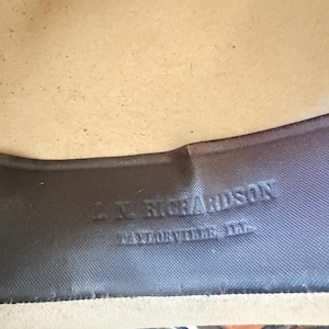 K&ouml;nnte beinhalten: Nahaufnahme des Inneren eines Hutes. Das Innenband des Hutes ist dunkelgrau mit gepr&auml;gtem Text, der "L.N. Richardson, Taylorville, Ill." lautet. Das Innere des Hutes ist hellbeige.