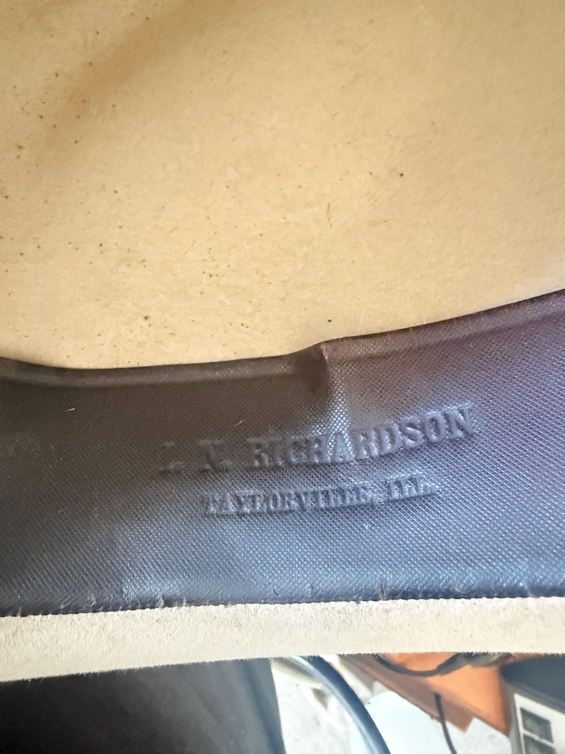 K&ouml;nnte beinhalten: Nahaufnahme des Inneren eines Hutes. Das Innenband des Hutes ist dunkelgrau mit gepr&auml;gtem Text, der "L.N. Richardson, Taylorville, Ill." lautet. Das Innere des Hutes ist hellbeige.