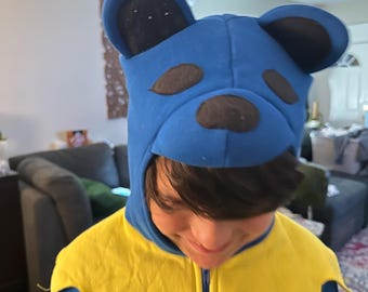 Puede incluir: Una sudadera con capucha azul con orejas de oso y un frente amarillo. La capucha tiene una cara de oso negro con una nariz marrón.