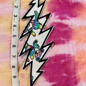 May include: A white lightning bolt patch with a black outline. The patch is decorated with tie-dye bears. The patch is approximately 6 inches long.
