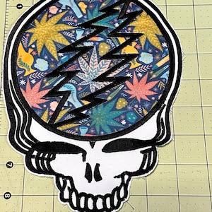 May include: A white fabric patch with a black outline of a skull and lightning bolt. The skull's eyes are black and the lightning bolt is filled with a colourful floral pattern. The pattern includes leaves, flowers, and other designs.