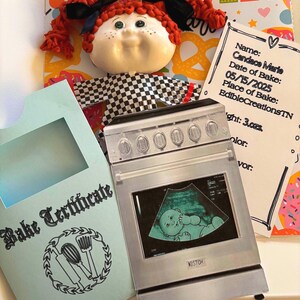 Może przedstawiać: Ręcznie robiona lalka z czerwonymi warkoczykami i czarną kokardą. Lalce towarzyszy miniaturowy piekarnik z obrazem USG. Obecny jest również "Bake Certificate" i karta z imieniem "Candace Marie".