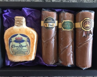 May include: A gift box containing a chocolate replica of a Crown Royal liquor bottle and three chocolate cigars. The cigars are wrapped in clear plastic and have labels that read "Happy Father's Day #1 Dad", "Dad Rocks", and a gold crown with a ribbon.
