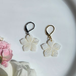 Może przedstawiać: Para białych kolczyków w kształcie kwiatów z rzeźbionym wzorem. Jeden kolczyk ma czarny haczyk, a drugi złoty.