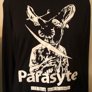 May include: Black long-sleeved top featuring a white graphic of a creature with large eyes and a blade. The word "Parasyte" is printed in white, with "-the maxim-" below it. A gold chain necklace is visible at the top.