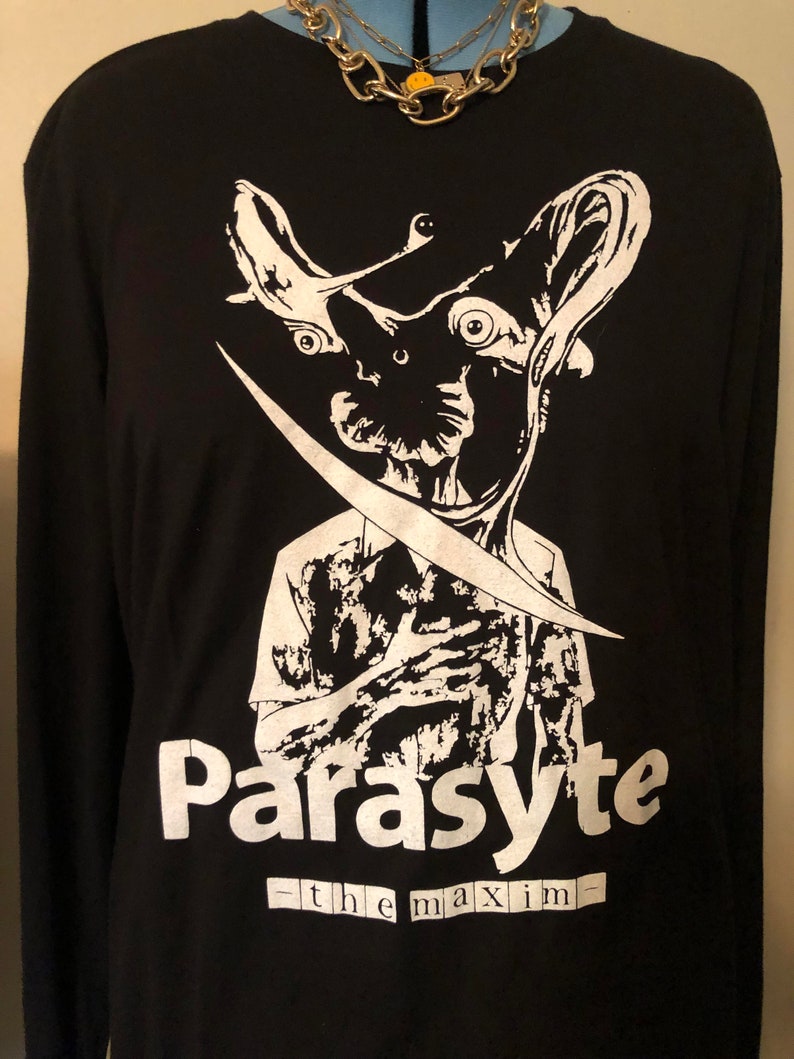 May include: Black long-sleeved top featuring a white graphic of a creature with large eyes and a blade. The word "Parasyte" is printed in white, with "-the maxim-" below it. A gold chain necklace is visible at the top.