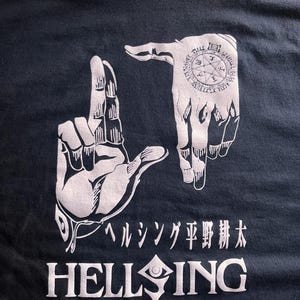 May include: Black t-shirt with a white graphic of two hands making the "L" sign. The hands have a pentagram symbol on the back. The text "HELLSING" is below the hands with a stylized eye and a drop of blood.
