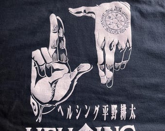 May include: Black t-shirt featuring a white graphic of two hands forming a frame, with a circular design on one hand. Japanese text and the word "HELLSING" are printed below the hands.