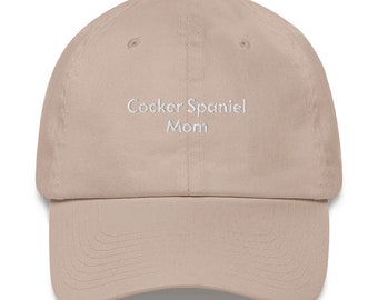 May include: A beige baseball cap with white embroidered text that reads "Cocker Spaniel Mom".