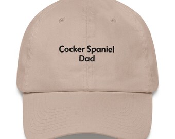 May include: A beige baseball cap with black embroidered text that reads "Cocker Spaniel Dad".