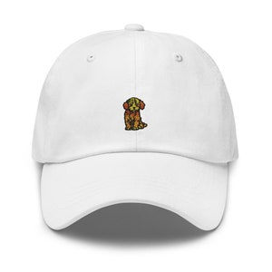 May include: A white baseball cap with a curved peak. The cap has a small embroidered image of a golden-coloured dog on the front. The dog is sitting, with its head and ears perked up. The cap has a classic six-panel design.