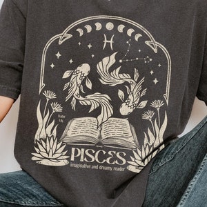 May include: A black t-shirt featuring a graphic of a book, two fish, and the zodiac symbol for Pisces. The text "Pisces" is below the graphic, along with the words "imaginative and dreamy reader".