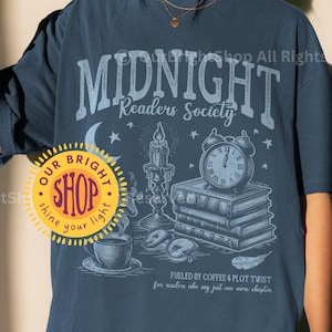 May include: Navy blue t-shirt with a graphic that reads "MIDNIGHT Readers Society." The design includes a crescent moon, stars, a candle, books, and an alarm clock. The text "FUELED BY COFFEE & PLOT TWIST" is also featured. A round shop logo is visible.