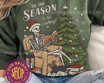May include: Military green sweatshirt with a festive graphic. The design features a skeleton wearing a Santa hat, reading a book near a Christmas tree. Text above reads "'Tis the Season To be reading!" A cozy and unique holiday themed top.