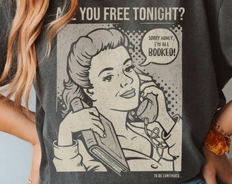 May include: A dark grey t-shirt featuring a vintage-style graphic of a woman on the phone. The graphic says "Are you free tonight?" and "Sorry honey, I'm all booked!"