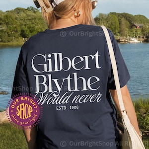 May include: A navy blue t-shirt featuring the white text "Gilbert Blythe Would never ESTD 1908". The shirt is worn outdoors near a lake. The shirt also has a small logo that says "Our Bright Shop".