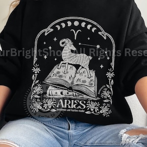 May include: Black sweatshirt with a white graphic design featuring an illustration of an Aries zodiac sign with a ram sitting on a stack of books. The design includes stars, moons, and floral elements. The text "ARIES" is included in the design.