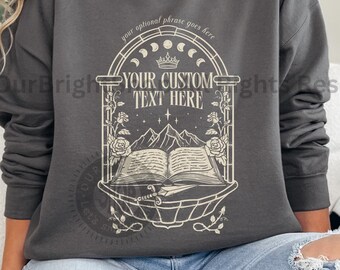Peut inclure: Un sweat-shirt gris foncé avec un motif de texte personnalisé. Le motif comprend un livre ouvert, des montagnes et des éléments floraux dans un cadre en arc. Le texte "YOUR CUSTOM TEXT HERE" est visible. Les options d'impression sont ivoire et charbon.