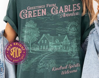 May include: A blue spruce t-shirt with a graphic that reads "Greetings From Green Gables Avonlea". The design includes an illustration of a house, tree, and the words "Kindred Spirits Welcome". The shirt also has a circular logo that says "Our Bright Shop".