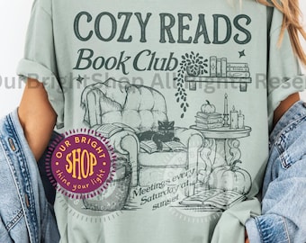 May include: Sage green t-shirt with a graphic that reads "COZY READS Book Club." The illustration depicts a cozy reading scene with a cat in a chair, books, and pumpkins. The shirt has a relaxed fit.