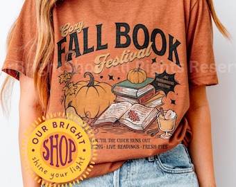 May include: Heather autumn-colored t-shirt with a graphic that reads "Cozy Fall Book Festival." The design includes pumpkins, books, and a beverage. The shirt is worn with light blue jeans.