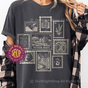 May include: Dark gray t-shirt featuring a collection of vintage-style postage stamps. The stamps depict various scenes, including a house, a gate, and botanical illustrations. The text "Wuthering Heights" is visible on one stamp. The shirt is labeled "PEPPER".