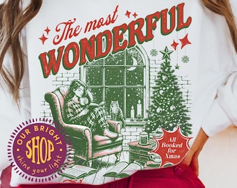 May include: White sweatshirt with a Christmas-themed graphic. The design features the text "The most WONDERFUL" in red and green, along with an illustration of a person reading by a window, a Christmas tree, and the text "All Booked for Xmas."