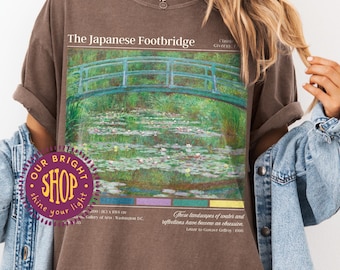 May include: Espresso-colored t-shirt featuring an image of "The Japanese Footbridge" with a green and blue bridge over a pond with lily pads. The shirt has text that reads "OUR BRIGHT SHOP" and "shine your light".