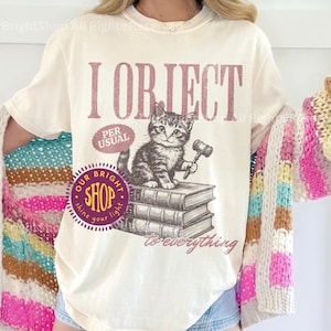 May include: Ivory-colored t-shirt with the text "I Object" and a kitten illustration holding a gavel. The shirt also features the text "Per Usual" and "to everything". The shirt is paired with a colorful striped cardigan.